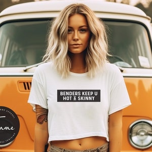 May include: A white cropped t-shirt with the text "BENDERS KEEP U HOT & SKINNY" printed in black on a black rectangle. The shirt is being worn by a person with blonde hair standing in front of a yellow vintage van.