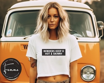 May include: A white cropped t-shirt with the text "BENDERS KEEP U HOT & SKINNY" printed in black on a black rectangle. The shirt is being worn by a person with blonde hair standing in front of a yellow vintage van.