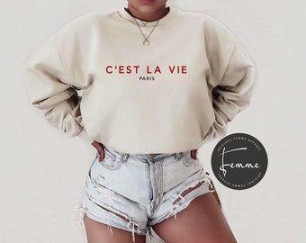 May include: A beige sweatshirt with the phrase "C'EST LA VIE PARIS" in red lettering. The sweatshirt is a relaxed fit and has long sleeves. The image also includes denim shorts.