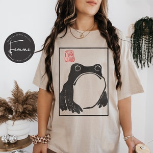 May include: A beige t-shirt with a black and white illustration of a frog. The frog is in a square frame with a red stamp in the top left corner. The text "Femme" is on a black circle in the top left corner of the image.