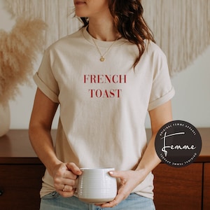 May include: A beige t-shirt with the text "FRENCH TOAST" in red lettering. The shirt is being worn by a person holding a white mug.
