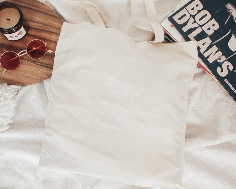 May include: A white canvas tote bag with a pair of round red sunglasses and a candle on a wooden cutting board. The tote bag is lying on a white blanket.