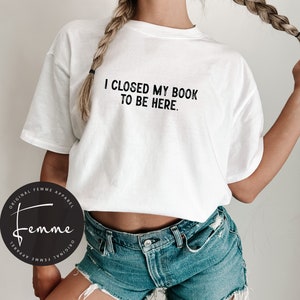 May include: White t-shirt with the text "I closed my book to be here." printed in black. The shirt is being worn by a person with long blonde hair. The person is wearing blue denim shorts.