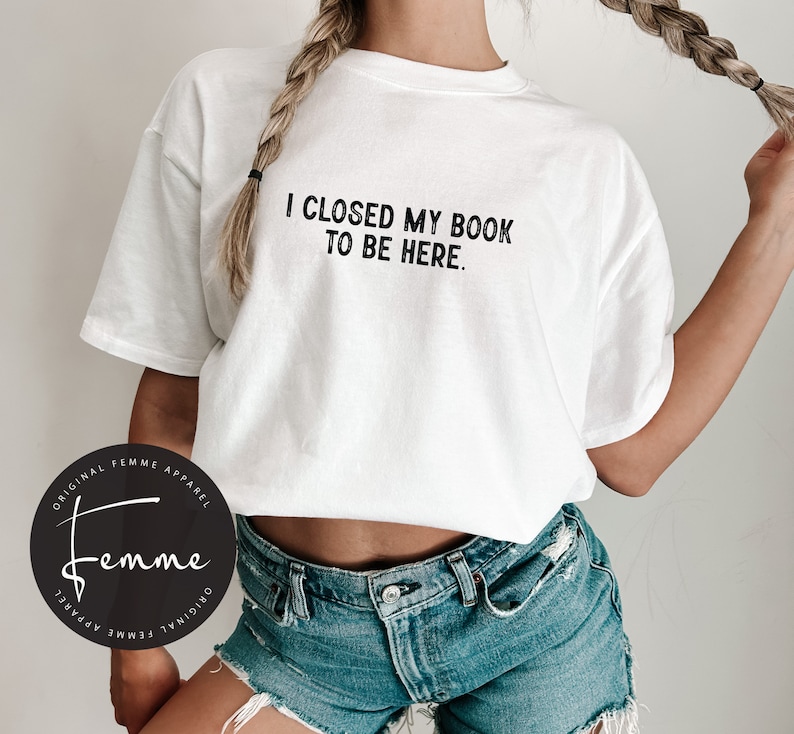 May include: White t-shirt with the text "I closed my book to be here." printed in black. The shirt is being worn by a person with long blonde hair. The person is wearing blue denim shorts.