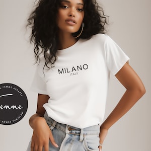 May include: White t-shirt with the text "MILANO ITALY" in black. The shirt is part of the "Femme" apparel line. The model is wearing blue jeans and gold hoop earrings.