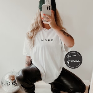 May include: A white t-shirt with the word "NOPE." printed in black on the front. The model is wearing black leather pants and a black and green trucker hat. The hat has a patch with a cartoon image of cowboy boots.
