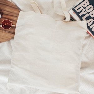 May include: A white canvas tote bag with a pair of round red sunglasses and a candle on a wooden cutting board. The tote bag is lying on a white blanket.