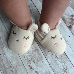May include: A pair of baby booties with a light brown and white striped pattern and a cute embroidered animal face with closed eyes. The booties are made of soft fabric and have a comfortable fit.