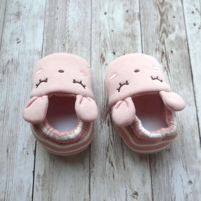 May include: A pair of pink baby booties with bunny ears and embroidered eyes. The booties have a white stripe around the top and bottom.