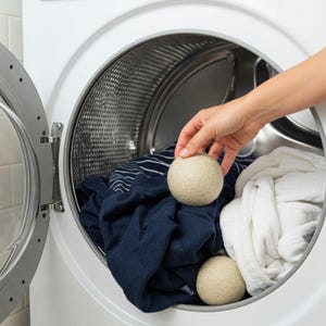 Peut inclure: Deux balles de s&eacute;chage en laine beige &agrave; l'int&eacute;rieur d'un tambour de machine &agrave; laver blanc avec une chemise ray&eacute;e bleu et blanc et une serviette blanche.