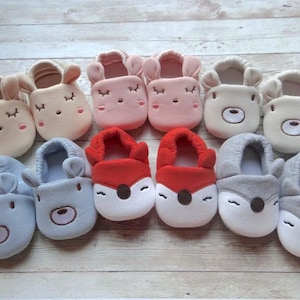 May include: Ten pairs of baby slippers with different animal designs. The slippers are made of soft, plush fabric and feature embroidered eyes, noses, and mouths. The colors include pink, red, blue, gray, and beige.
