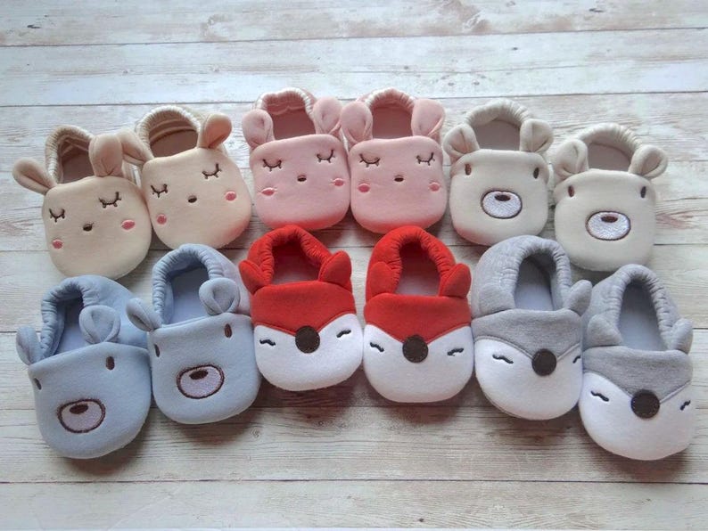 May include: Ten pairs of baby slippers with different animal designs. The slippers are made of soft, plush fabric and feature embroidered eyes, noses, and mouths. The colors include pink, red, blue, gray, and beige.