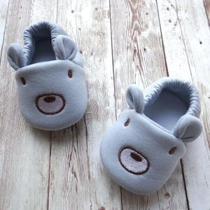May include: A pair of light blue baby slippers with a bear face design. The slippers have brown embroidered eyes, nose, and mouth.