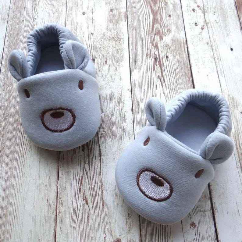 May include: A pair of light blue baby slippers with a bear face design. The slippers have brown embroidered eyes, nose, and mouth.