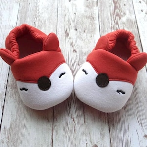 May include: A pair of red and white fox-shaped baby slippers with black eyes and a brown nose.