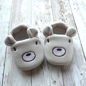 May include: A pair of beige baby booties with embroidered bear faces. The booties have brown stitching around the bear's eyes and nose.