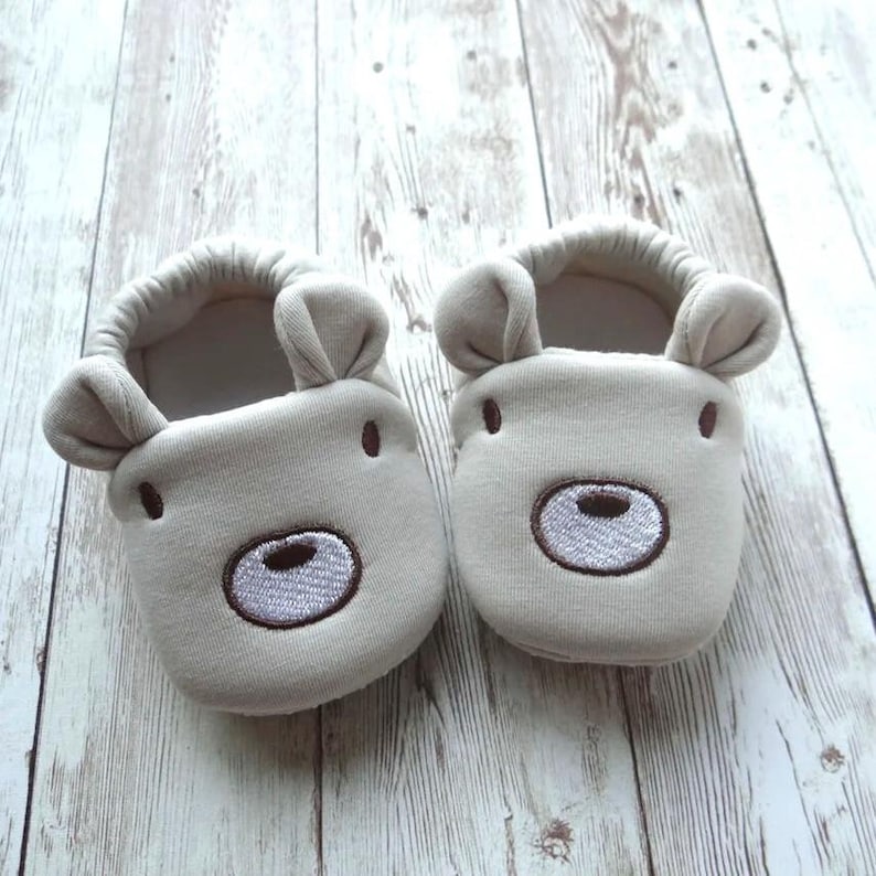 May include: A pair of beige baby booties with embroidered bear faces. The booties have brown stitching around the bear's eyes and nose.