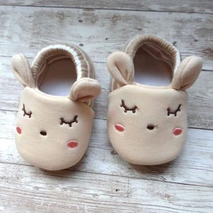 May include: A pair of beige baby slippers with embroidered eyes and a nose, shaped like a bunny with floppy ears.