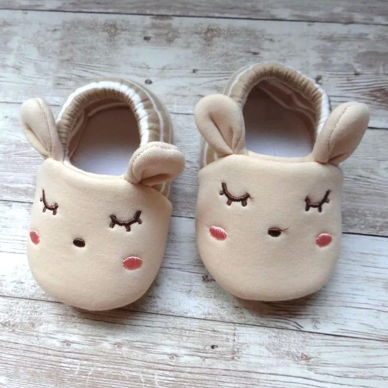 May include: A pair of beige baby slippers with embroidered eyes and a nose, shaped like a bunny with floppy ears.