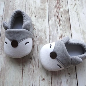 May include: A pair of gray and white baby slippers with a fox design. The slippers have a gray fabric upper with white accents and a brown nose.