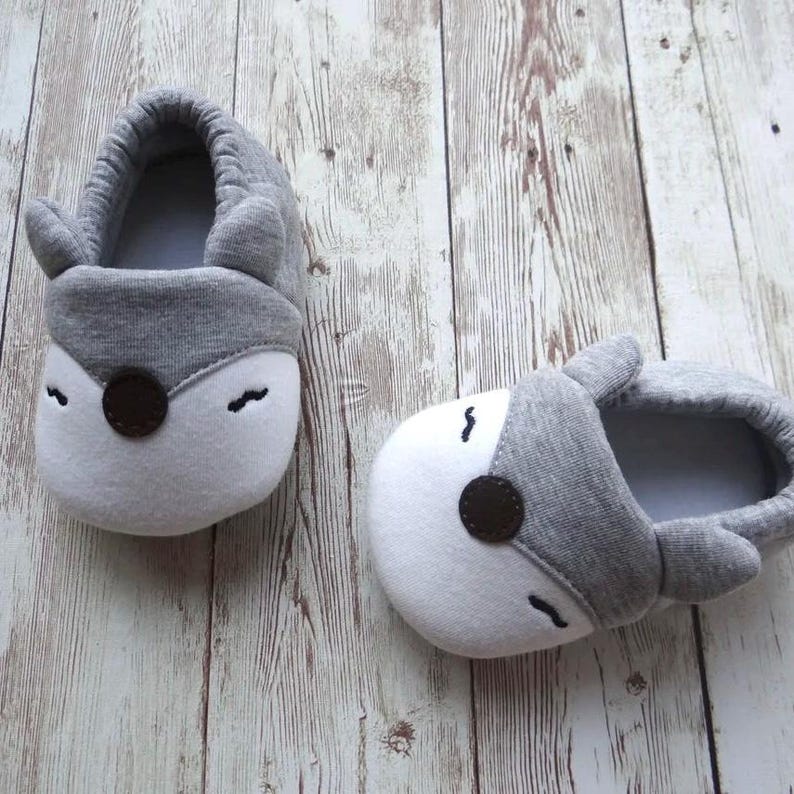 May include: A pair of gray and white baby slippers with a fox design. The slippers have a gray fabric upper with white accents and a brown nose.