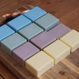 May include: A set of 12 square-shaped soap bars in various pastel colors, including light blue, lavender, yellow, and green. The soap bars are arranged in a grid pattern on a wooden surface.