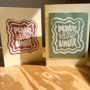 以下が含まれることがあります： 緑と茶色の背景に白い文字で「Healing is not linear」と書かれたカードが2枚。カードは木製の表面の上に置かれています。