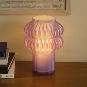 May include: A lavender-colored table lamp with a ribbed base and a stacked, rounded shade. The lamp is lit, emitting a warm glow. The lamp is on a wooden surface.