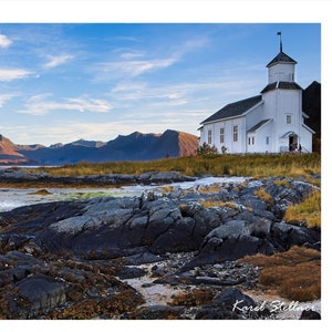 May include: A white church with a steeple stands on a rocky shore, overlooking a body of water with mountains in the background. The sky is blue with white clouds.