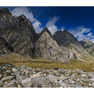 Może przedstawiać: Panoramiczny widok na skalisty pasmo górskie z błękitnym niebem i białymi chmurami. Góry są szare i brązowe z zielonym, trawiastym obszarem na pierwszym planie. W oddali widać kilka małych czerwonych namiotów.
