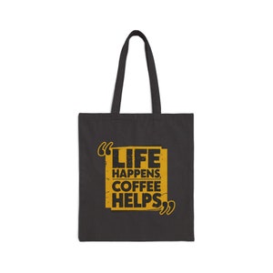 以下が含まれることがあります： 黒いトートバッグに、黄色い長方形の中に黒い文字で「Life Happens, Coffee Helps」と引用符が書かれています。