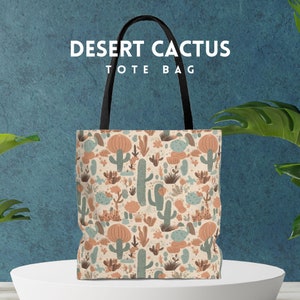 May include: A beige tote bag with a black handle. The bag has a repeating pattern of cacti and flowers in shades of green, brown, and orange. The text "DESERT CACTUS TOTE BAG" is printed on the bag.