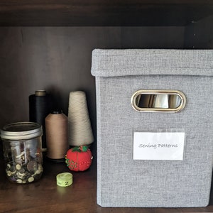 May include: A gray fabric storage box with a silver metal handle and a label that says "Sewing Patterns". The box is sitting on a shelf with various spools of thread and a jar of buttons.
