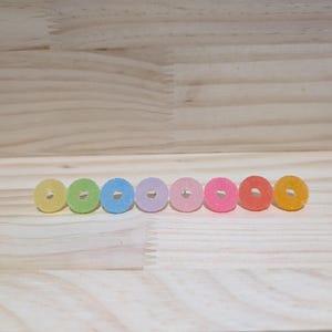 May include: Seven colourful, ring-shaped sweets arranged in a row. The sweets are in a range of colours: yellow, green, blue, lilac, pink, red-orange, and orange. Each sweet has a hole in the middle and a slightly frosted appearance.