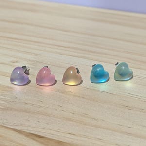 May include: Five heart-shaped stud earrings in a row, each with a different translucent color: purple, pink, orange, blue, and green. The earrings are made of a glossy material and have silver posts.