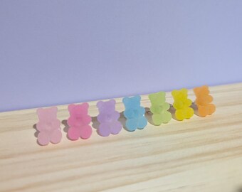 May include: A collection of colorful gummy bear-shaped stud earrings in a row. The earrings are in shades of pink, purple, blue, green, yellow, and orange. They are arranged on a light wooden surface against a purple background.