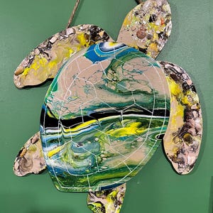 May include: A wall-hanging sea turtle cutout featuring a fluid acrylic pour design in shades of blue, green, yellow, black, and white. The shell is detailed with white painted geometric lines and the flippers are accented with small embedded seashells.