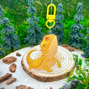 May include: A yellow bearded dragon keychain with a yellow plastic clasp. The keychain is made of clear acrylic and features a cartoon-style illustration of a bearded dragon.