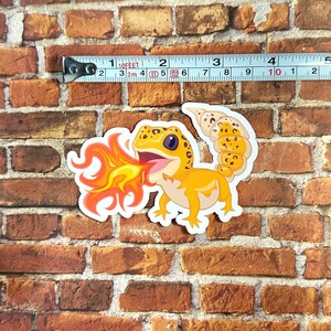 Peut inclure: Un autocollant repr&eacute;sentant un gecko de dessin anim&eacute; avec des &eacute;cailles orange et jaunes crachant du feu. Le gecko est sur un fond de briques.