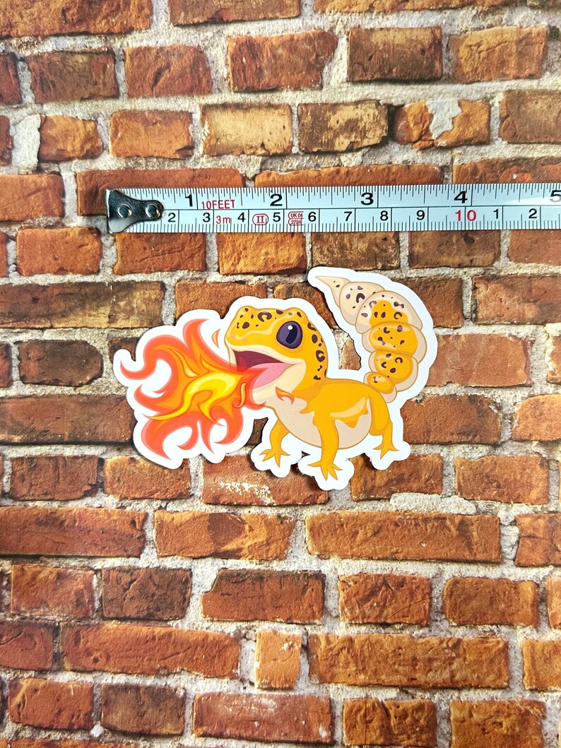Peut inclure: Un autocollant repr&eacute;sentant un gecko de dessin anim&eacute; avec des &eacute;cailles orange et jaunes crachant du feu. Le gecko est sur un fond de briques.