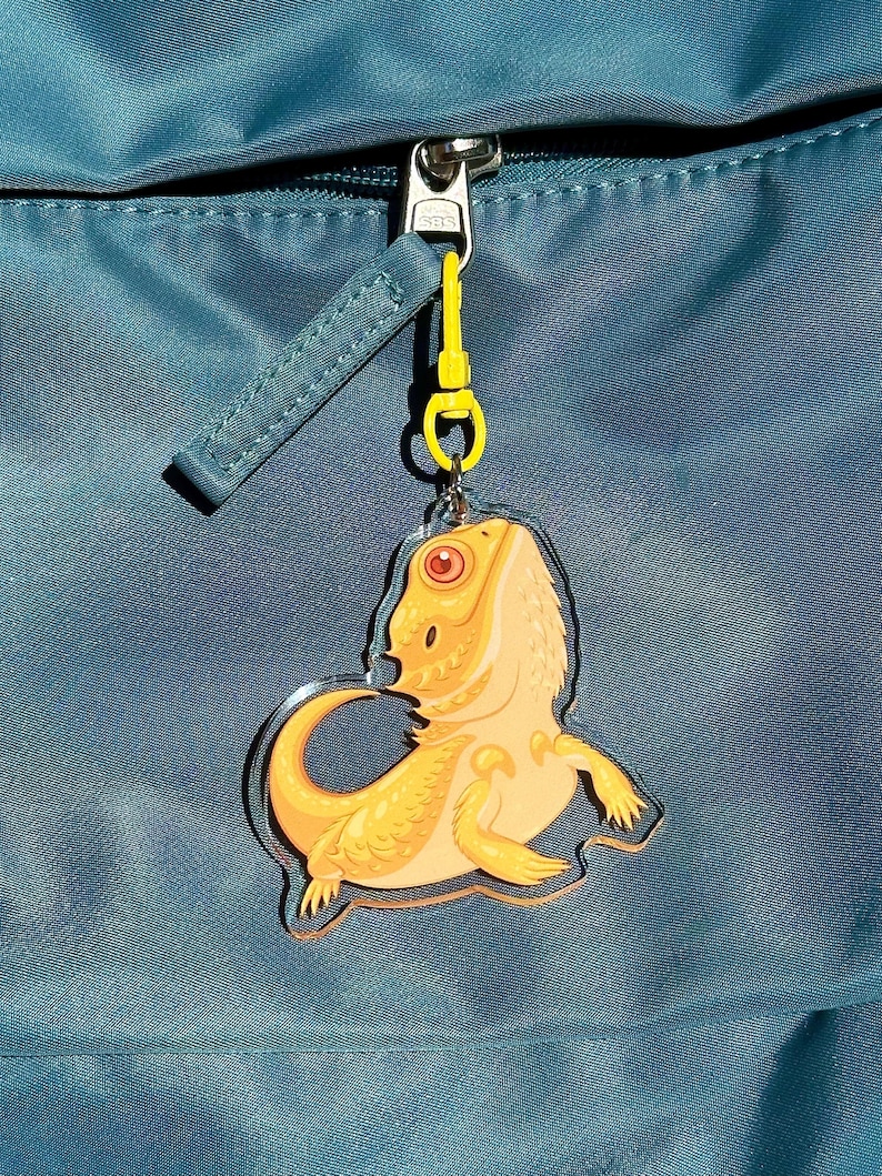 May include: A yellow bearded dragon keychain charm attached to a blue backpack zipper.