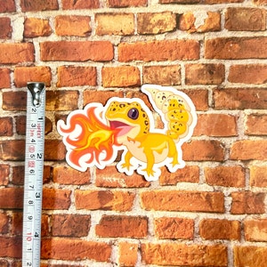Peut inclure: Un autocollant repr&eacute;sentant un gecko de dessin anim&eacute; avec des &eacute;cailles orange et jaunes crachant du feu. Le gecko est sur un fond de briques.