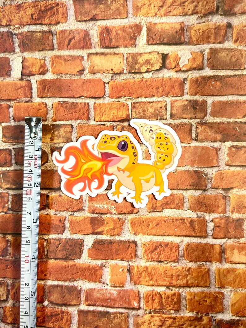 Peut inclure: Un autocollant repr&eacute;sentant un gecko de dessin anim&eacute; avec des &eacute;cailles orange et jaunes crachant du feu. Le gecko est sur un fond de briques.
