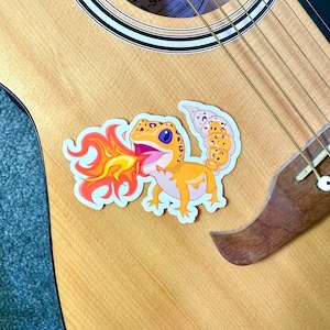 Peut inclure: Un autocollant repr&eacute;sentant un gecko de dessin anim&eacute; crachant du feu sur une guitare acoustique en bois. La guitare porte une &eacute;tiquette Fender A-Series sur le trou sonore.