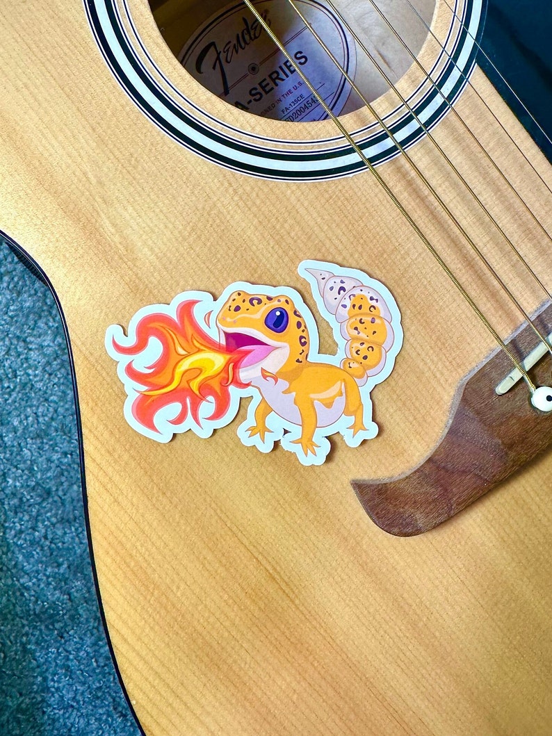 Peut inclure: Un autocollant repr&eacute;sentant un gecko de dessin anim&eacute; crachant du feu sur une guitare acoustique en bois. La guitare porte une &eacute;tiquette Fender A-Series sur le trou sonore.