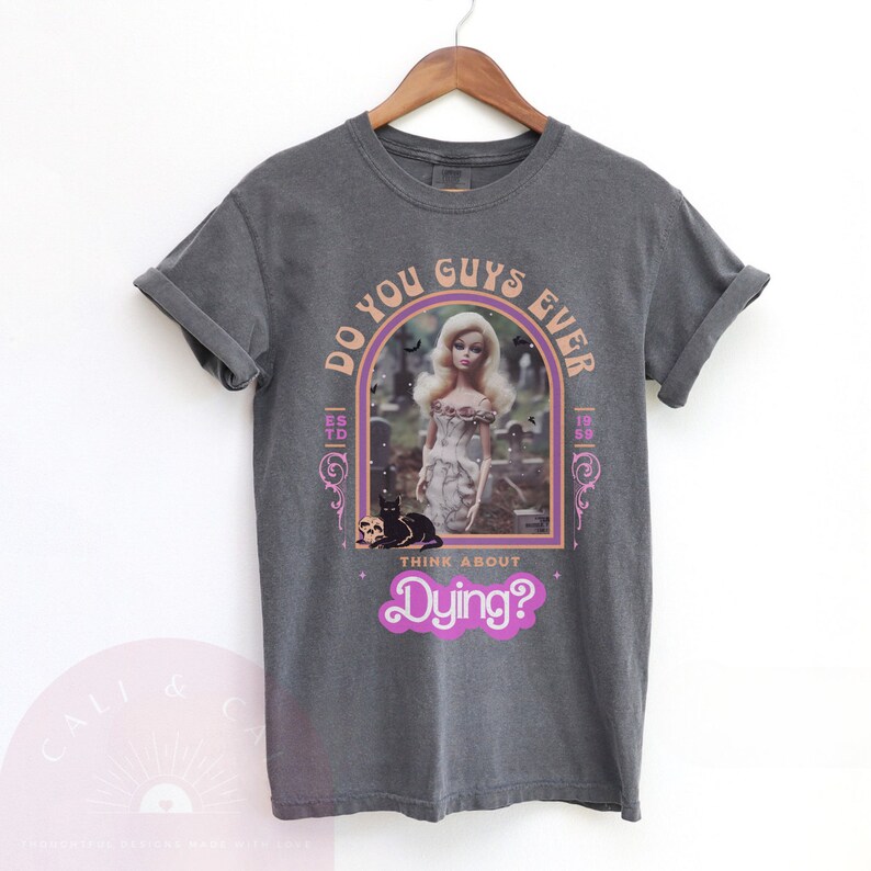 May include: A charcoal gray short-sleeve t-shirt featuring a graphic print of a blonde doll in a graveyard with a black cat. The text reads DO YOU GUYS EVER THINK ABOUT Dying? with ESTD 1959 on the sides, framed in a purple and orange arch.