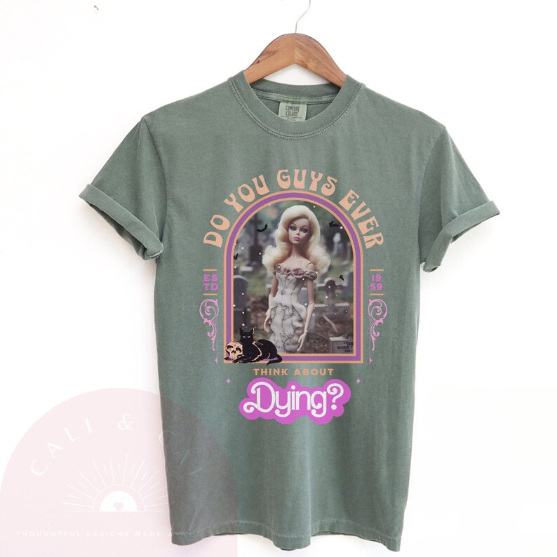 May include: A sage green short-sleeve t-shirt featuring a graphic print of a blonde doll in a graveyard with a black cat and skull. The text reads DO YOU GUYS EVER THINK ABOUT Dying? with ESTD 1959 on the sides, framed in an arch design.