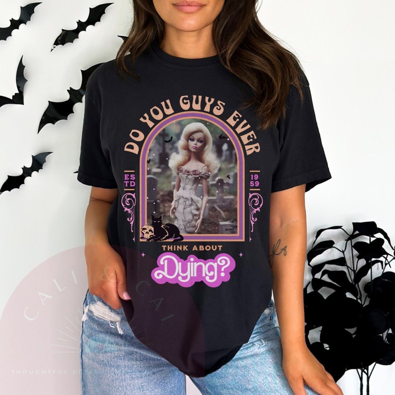 May include: A black t-shirt featuring a graphic print of a blonde doll in a graveyard with a black cat and skull. The text reads DO YOU GUYS EVER THINK ABOUT Dying? with ESTD 1959 on the sides. The design is framed by a purple and orange arch.