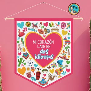 以下が含まれることがあります： 「MI CORAZÓN LATE EN dos idiomas」のテキストがハート型で書かれたピンク色のペナントバナー。文化的なアイコンのカラフルなイラストで囲まれています。バナーは木の棒から吊り下げられています。両側に2つのサボテンがあります。