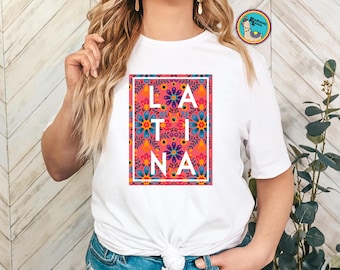 May include: White t-shirt featuring the word "LATINA" in white block letters, set against a vibrant floral pattern in a rectangular frame. The floral design includes blue, orange, and pink flowers. The shirt is tied at the waist.